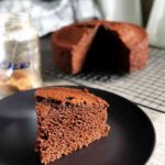 Fatia de bolo de chocolate servida sobre um prato preto com o bolo em segundo plano